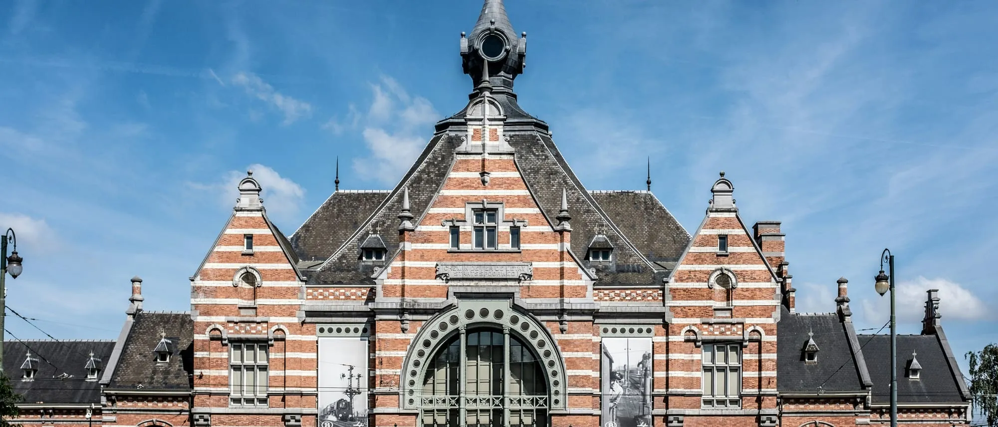 La façade en briques rouges et blanches de la gare de Schaerbeek sous un ciel bleu