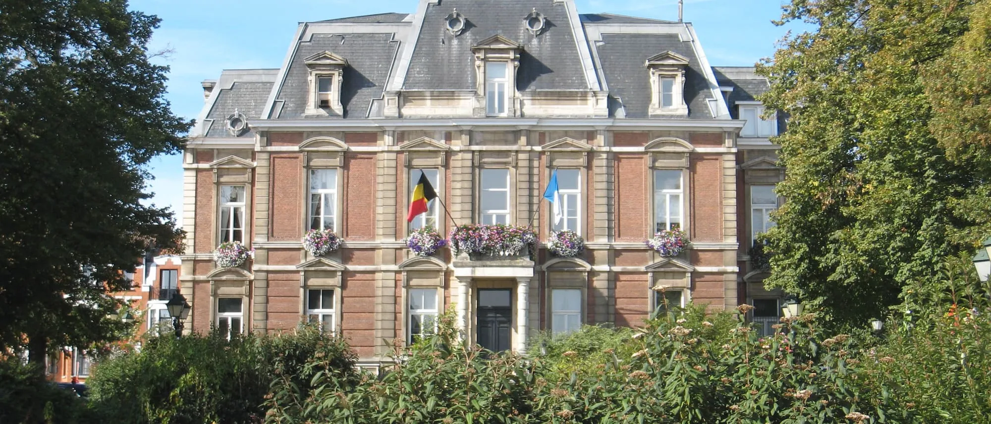 La maison communale d'Uccle, façade en briques et fenêtres fleuries, entourée de verdure